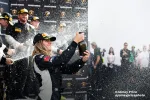 Race car driver celebrates with champagne spray on podium after winning motorsport event.
