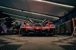 Front view of a red race car with open doors in a dimly lit garage surrounded by tools and crew members.