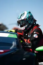 Race car driver in teal helmet and black suit leans on green and black race car on a clear day