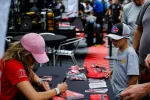 Young boy receives autograph from woman in pink cap at event table with racing merchandise and fans.