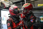 Two race car drivers in red and black suits and helmets share a light moment in a garage.