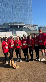 Group of seven runners in red shirts posing with medals after a race in front of a modern glass building on a sunny day.