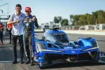 Two men celebrating next to a blue Acura Konica Minolta race car on a sunny racetrack.