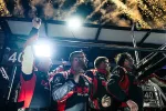Racing team in matching red and black suits celebrating with fireworks in the background at night.