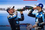 Two race car drivers celebrating with champagne on podium wearing racing suits and caps after a victory.