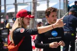 Woman in red cap gestures while talking to man in black Konica Minolta shirt at motorsport event.
