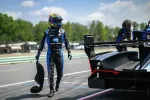 Race car driver in blue and black suit walking on track beside a black racing car with large rear wing.