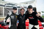 Two race car drivers and a team member posing in Victory Lane with a red race car and winner's board behind them