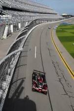 Red and black race car speeding on a curved asphalt track with empty grandstands at a professional racing circuit.
