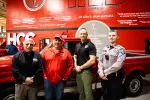 Four men, including police officers and a man in red, standing in front of a red truck and a red wall with quotes and HCC logos.