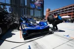 Blue Acura race car in pit stop with crew member holding tire, set against urban skyline on sunny day.
