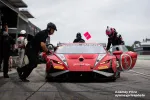 Red Lamborghini Huracan race car in pit stop with driver entering and crew members preparing under overcast sky.