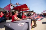 Racing team members in red shirts signing posters for fans at an outdoor motorsports event under clear blue sky.