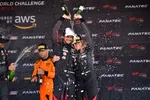 Race winners celebrate on podium spraying champagne with team in black and orange racing suits and caps