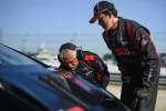 Two racing crew members in black uniforms inspecting a race car on track during daytime pit stop.