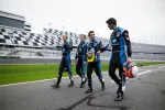 Four race car drivers in blue racing suits walking on a racetrack with helmets in hand near grandstands.