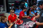 Race drivers and team members sign autographs for fans at Acura Motorsports event with merchandise on table.