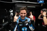 Excited race car driver in blue and black racing suit celebrating with both index fingers raised inside a garage.