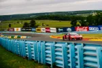 Red race car speeding on curved racetrack with blue barriers and sponsor banners in rural setting under cloudy sky