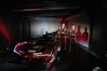 Two race car drivers in black and red suits stand beside a red Acura DEX race car in a dimly lit garage.