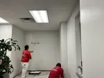 Two painters in red shirts applying white paint on office walls with rollers near a large green plant and ceiling lights.