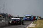 Black race car leads a pack of colorful sports cars on a racetrack under overcast sky