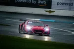 Red Acura race car with headlights on speeding on wet racetrack during a cloudy day race event.