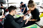 Man signing autograph for baby held by smiling woman at outdoor event with people in the background.