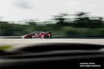 Red race car number 25 speeding on a blurred racetrack with a green forest background and overcast sky.