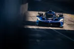 Blue Acura race car with headlights on driving on a sunlit race track pit lane.