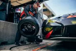 Race car mechanic in a black and orange suit changes tire on a sleek black race car during pit stop.