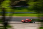 Red racing car speeding on a racetrack with blurred green trees and asphalt background.
