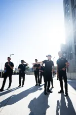 Group of seven men in black uniforms standing and talking outdoors on a sunny day casting long shadows.