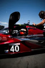Race car driver in red and black Acura prototype car number 40 during pit stop with crew member wearing headset.