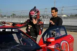 Race car driver in black and red suit adjusts helmet beside red Acura race car at pit stop with crew member helping.