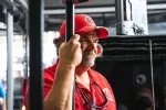 Man in red uniform and cap holding a black pole, looking focused with sunglasses hanging on his shirt.