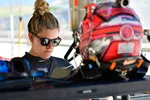 Female race car driver wearing sunglasses and reviewing notes next to a red racing helmet.