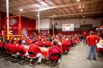 Large group of workers in red uniforms attending safety training session in a spacious industrial warehouse.