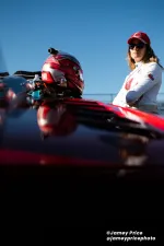 Female race car driver in white suit and red hat stands with arms crossed by red racing car and helmet.