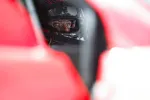 Race car driver wearing helmet and glasses, seen through a red car window, focused and ready to drive.