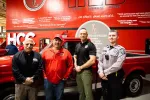 Four men including a police officer and workers stand in front of a red truck inside a facility with red walls and HCC branding.