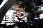 Mechanic wearing sunglasses working on car engine with focus on wiring and dashboard components.