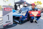 Race car driver kneels beside blue Acura race car holding Motul Pole Award banner next to number 1 GTP sign.