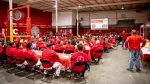 Workers wearing red shirts attend a safety meeting in a large industrial warehouse with red and white decor.