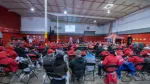 Large group of people in red jackets attending a meeting in a spacious indoor hall with chairs and projector screen.