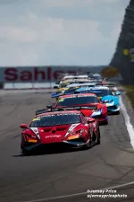Red Lamborghini leading a pack of racing cars on a curving racetrack under a cloudy sky.