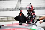 Race car driver wearing a pink and black helmet and suit climbs into a race car during a pit stop.