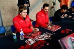 Two racecar drivers in red jackets signing posters and cards at an autograph table with fans nearby.