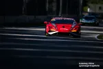 Red race car with sponsor logos speeding on a shaded racetrack with another car in the background.
