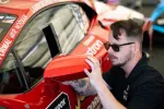 Man wearing sunglasses adjusts side mirror on red race car with sponsor decals in garage setting.